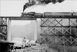 A steam locomotive pulling passenger cars crosses a bridge under which onlookers stand next to mid-twentieth-century automobiles.